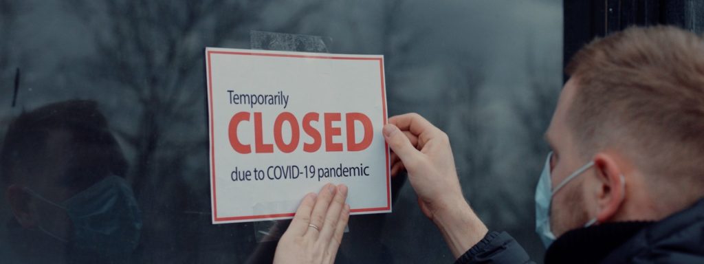 Mann klebt ein Schild mit Text "Geschlossen wegen der COVID-19 Pandemie" an eine Fensterscheibe